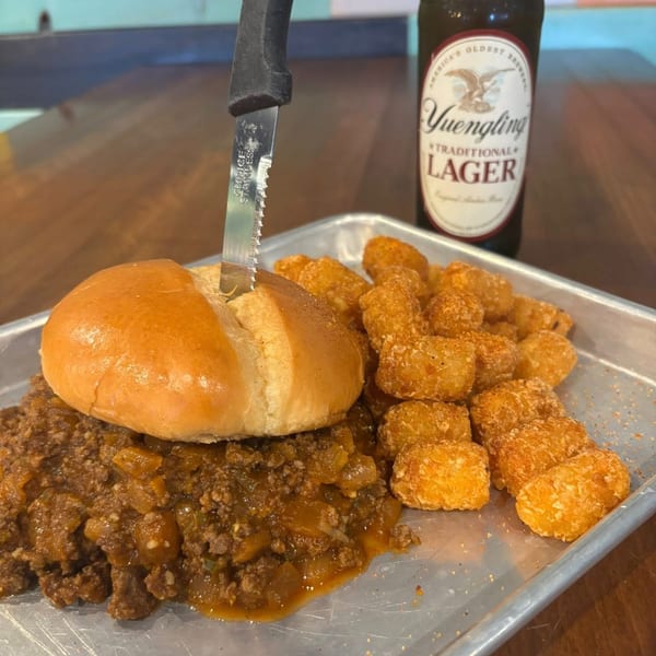 A chili cheeseburger with a knife, tater tots, and a Yuengling beer on a metal tray.