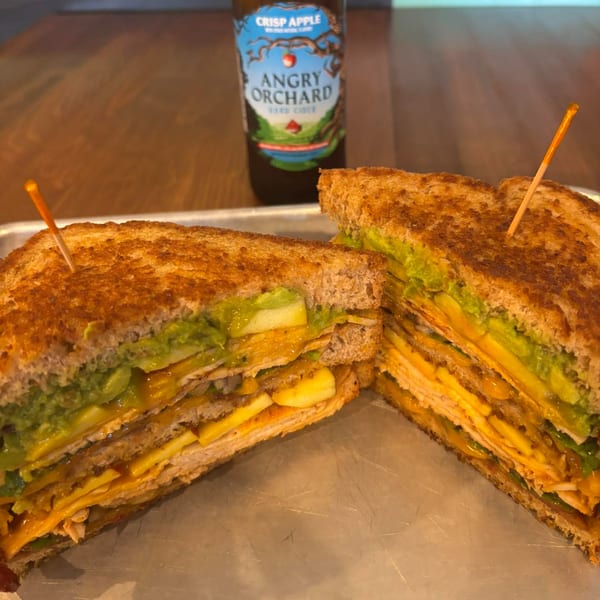 A grilled sandwich with avocado, cheese, and meat, cut in half, with a bottle of Angry Orchard cider.