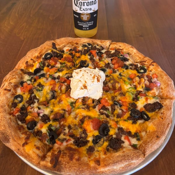 A taco pizza with ground beef, olives, tomatoes, cheese, and a dollop of sour cream, with a Corona beer bottle in the background.