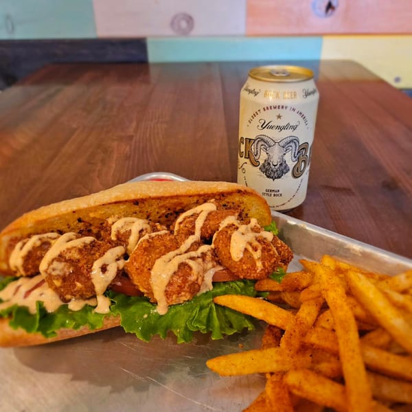 A fried shrimp po'boy with lettuce, tomato, and sauce, served with fries and a Yuengling beer.